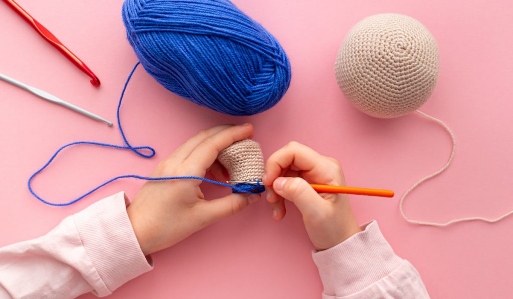 Children's hands in the process of crocheting toys from blue and beige yarn.
