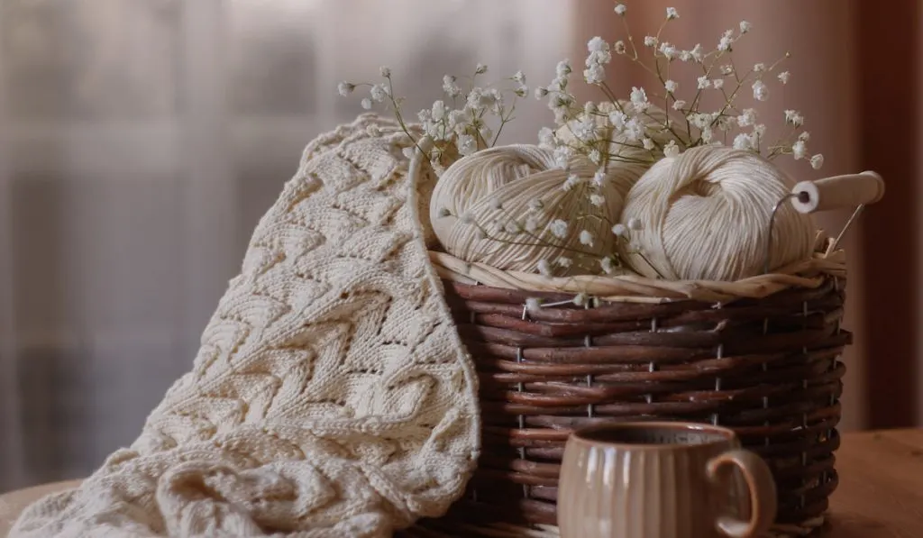Home cozy wicker basket with yarn, knitted blanket, knitting needles and a cup of tea.