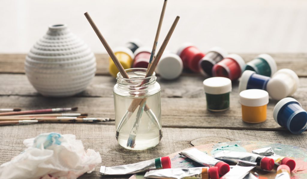 Paint brushes in a solvent and scattered containers with poster paints and acrylic paints tubes
