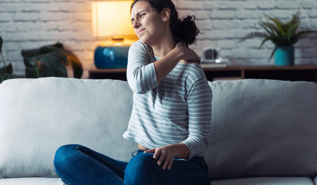 Young woman experiencing shoulder pain sitting on the sofa in the living room 