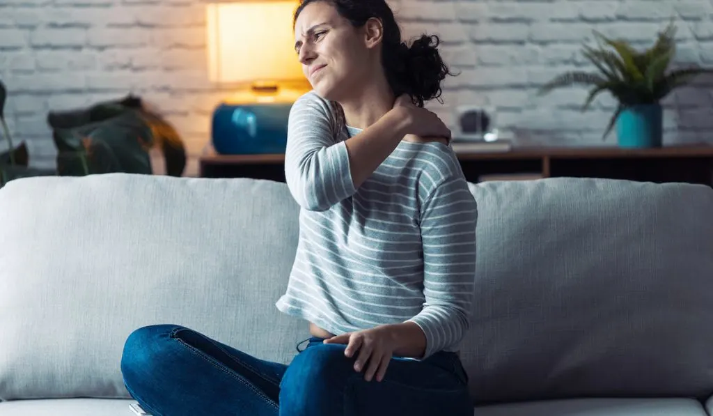 Young woman experiencing shoulder pain sitting on the sofa in the living room