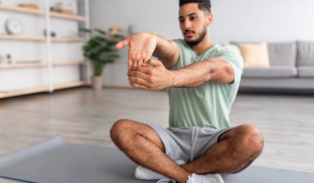 man sitting on sports mat in lotus pose, stretching hands at home