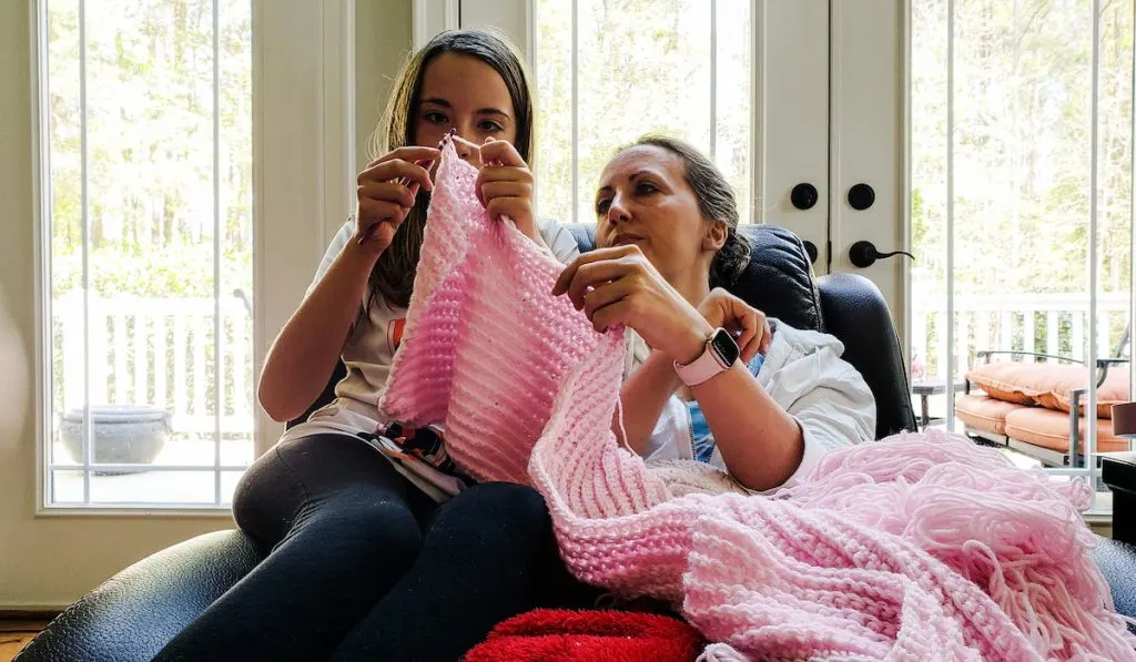 mother and daughter helping each other to make knitted blanket using pink yarn 