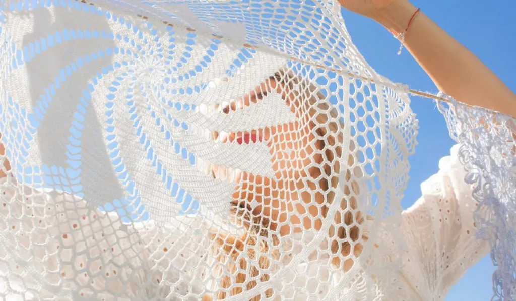 woman hanging clean white laundry on rope and looking through knitted doily