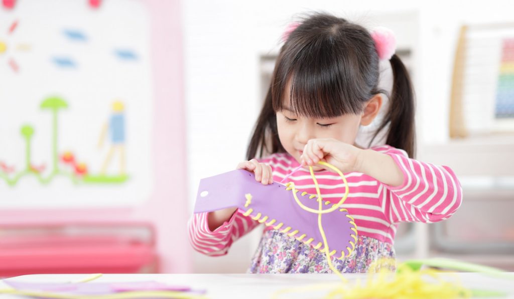Young adorable girl in pink dresss sewing pad craft using plastic needle and yarn for homeschooling