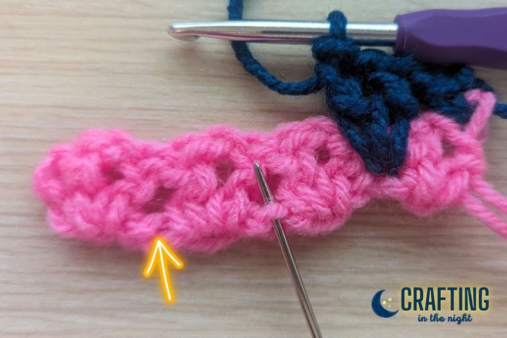 crochet swatch showing first stitches of row 2 complete and a yarn needle indicating where the next stitch should go. A yellow arrow indicates where the subsequent stitch should be placed