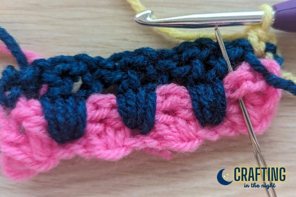 a crochet swatch showing where the first v-stitch of the pattern should be worked for row 3. The space is indicated with a yarn needle.