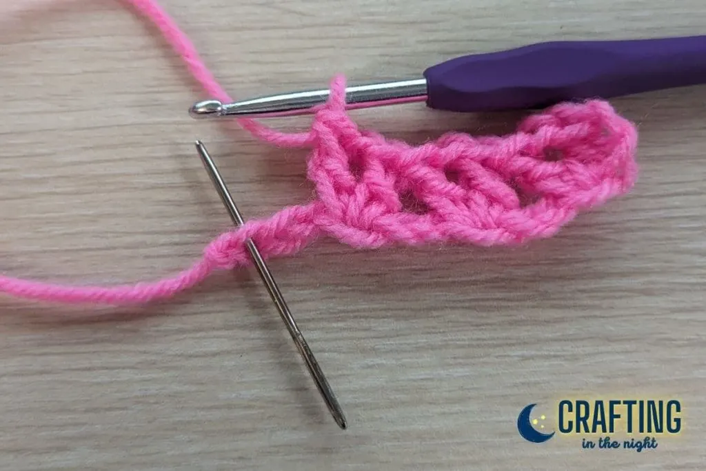 A crochet swatch with three v-stitches. A yarn needle is placed through the last chain on the foundation row to indicate where the last v-stitch should be placed.