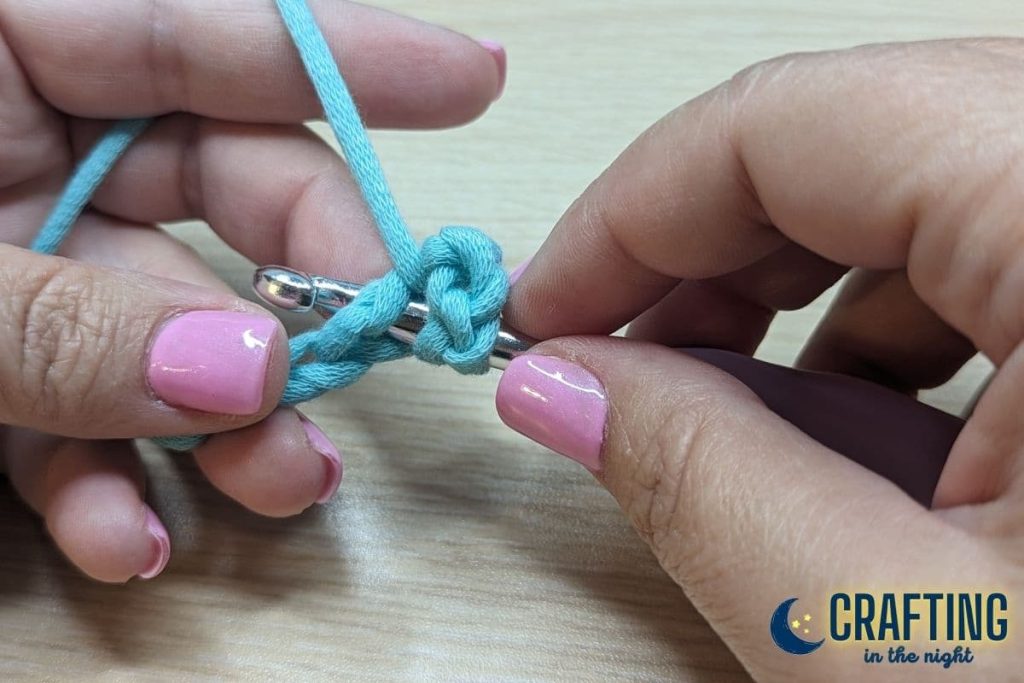 A chain of blue yarn held between two hands. A metal crochet hook, inserting the hook under the fourth stitch from the hook, skipping three chains.