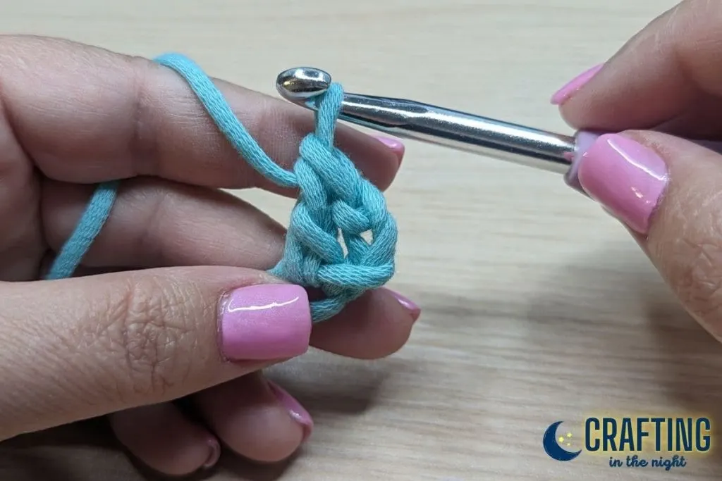 A chain of blue yarn held between two hands. A metal crochet hook, pulling the yarn through the remaining two loops on the hook. Completed one double crochet stitch.