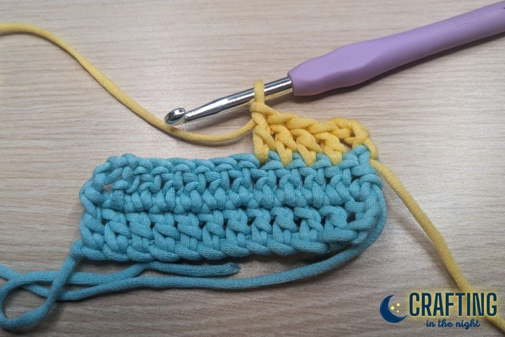 A double crochet of blue yarn with a few yellow yarn stitches with a crochet hook on the table.