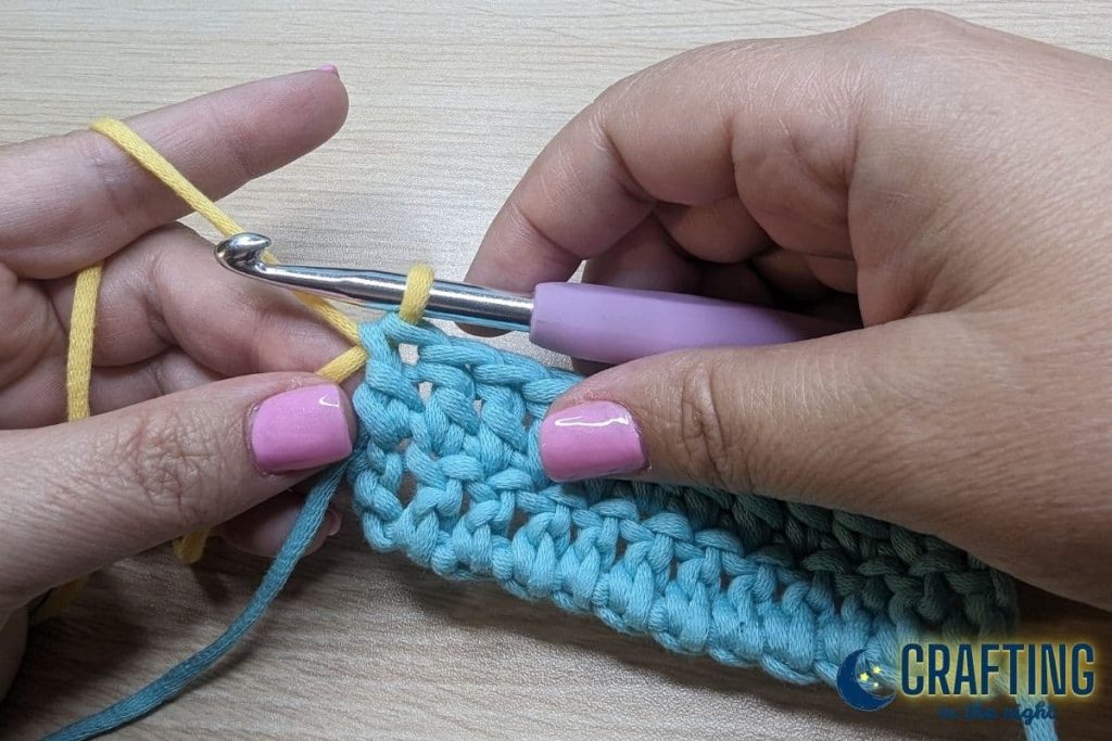 A double crochet of blue yarn and a metal crochet hook. Yarn over with the yellow yarn and pull it through the two loops left on the hook.