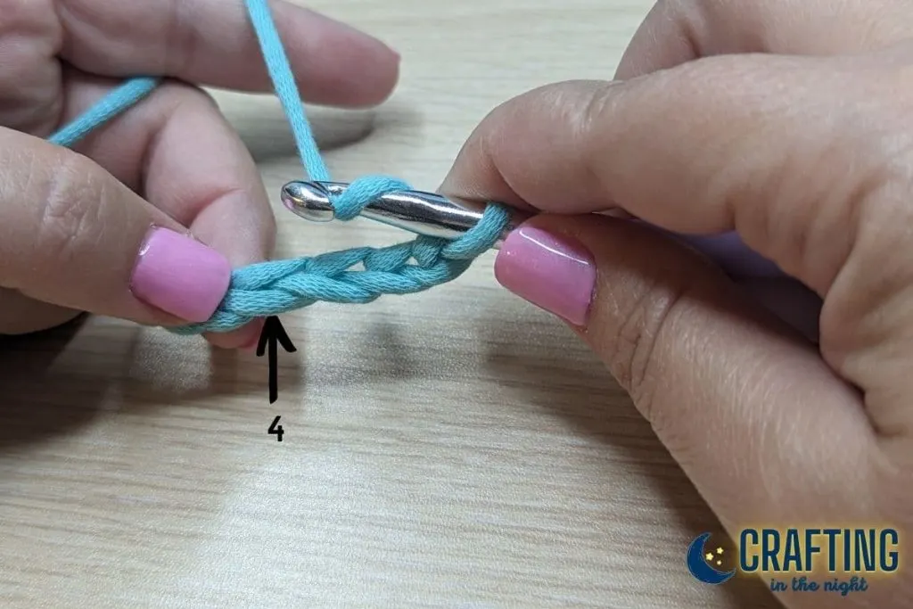 A chain of blue yarn held between two hands. A metal crochet hook has a loop on the hook and yarn wrapped over it from back to front. A black arrow indicates the fourth stitch from the hook.