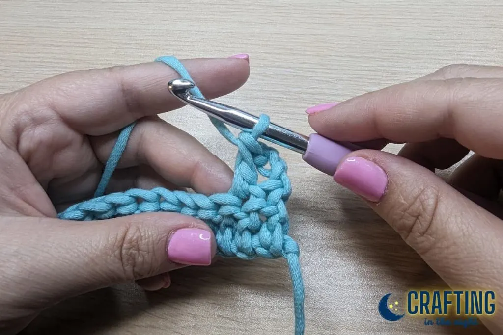 A chain of blue yarn held between on the other hand and a crochet hook on the other hand. Skip the first stitch and place a double crochet into the next stitch. 