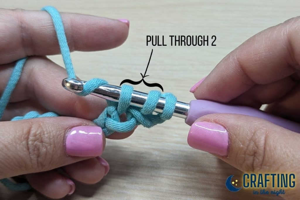 A chain of blue yarn held between two hands. A metal crochet hook pulling the yarn through the first two loops on the hook. A black arrow indicates the first two loops from the hook.