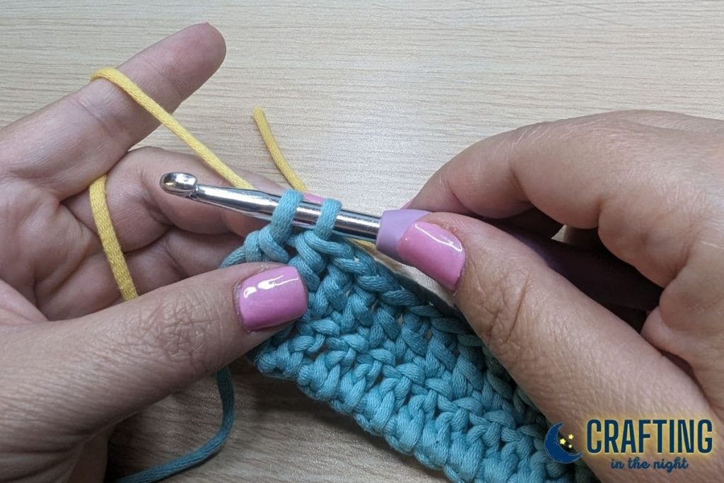 A double crochet of blue yarn and a metal crochet hook. A yellow yarn positioned over the left pointing finger. Setting up the new yarn color.