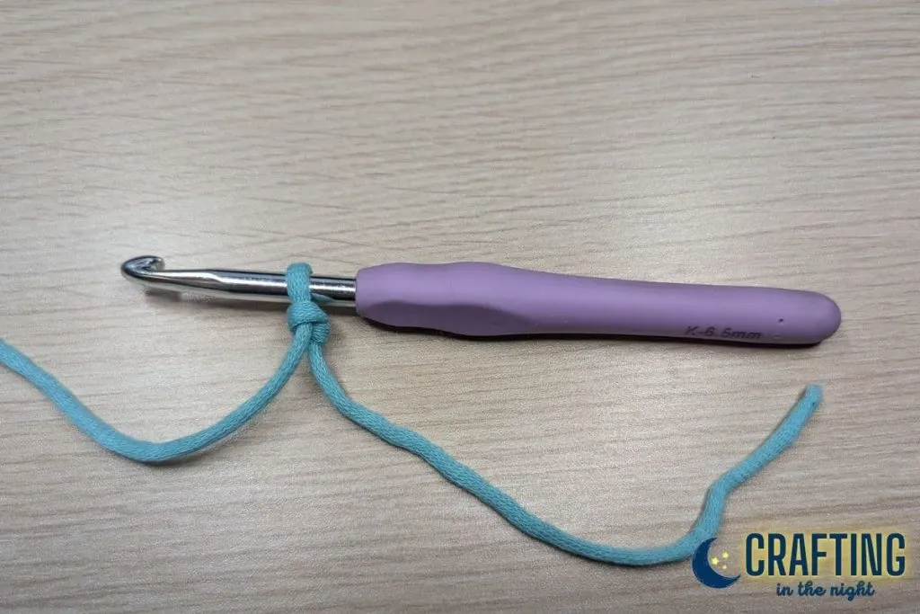 Blue yarn forms a slip stitch on a crochet hook with a purple handle.