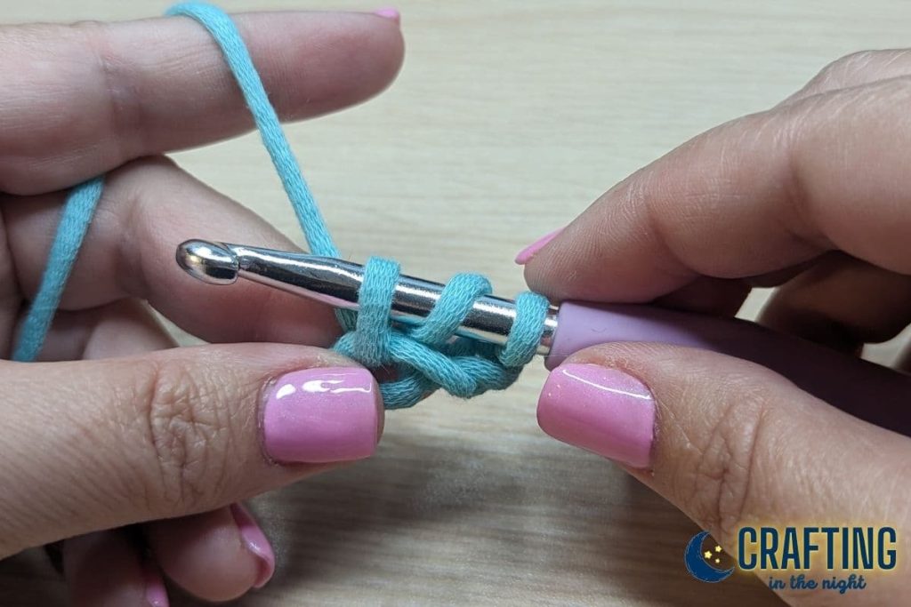 A chain of blue yarn held between two hands. A metal crochet hook, pulling the yarn through the stitch. There are three loops on the hook.