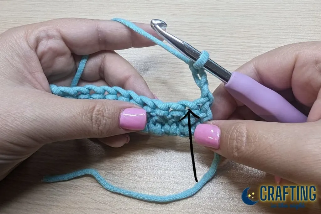 A chain of blue yarn held between two hands. Turn your work so that you are looking at the opposite side. A black arrow showing the opposite side.