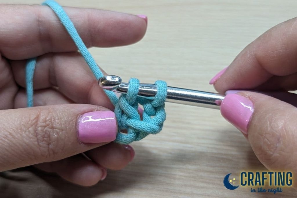 A chain of blue yarn held between two hands. A metal crochet hook leaving two loops on the hook.