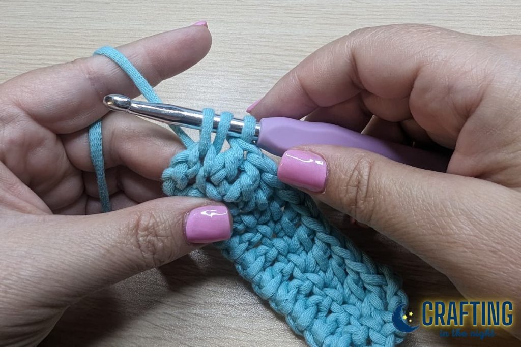A chain of blue yarn yarn held between two hands and a metal crochet hook. Place your last double crochet into the top of the chain 3 from the previous row.