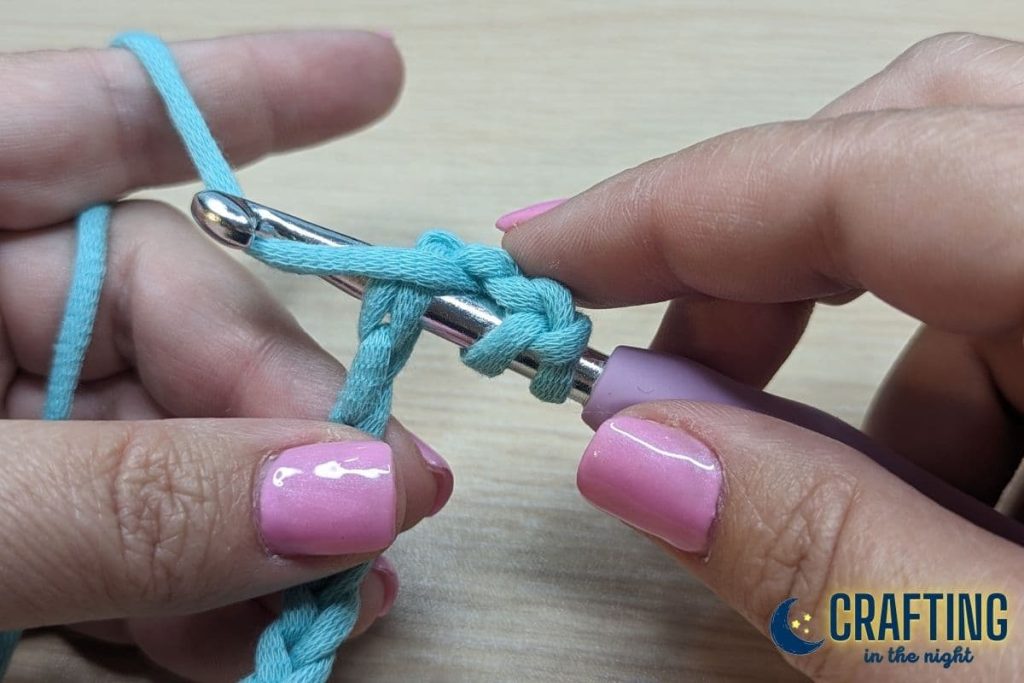 A chain of blue yarn held between two hands. A metal crochet hook, yarn over the hook again.
