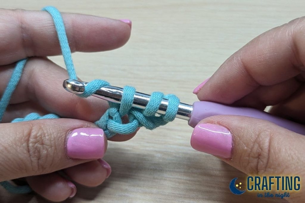 A chain of blue yarn held between two hands. A metal crochet hook, yarn over the hook again.