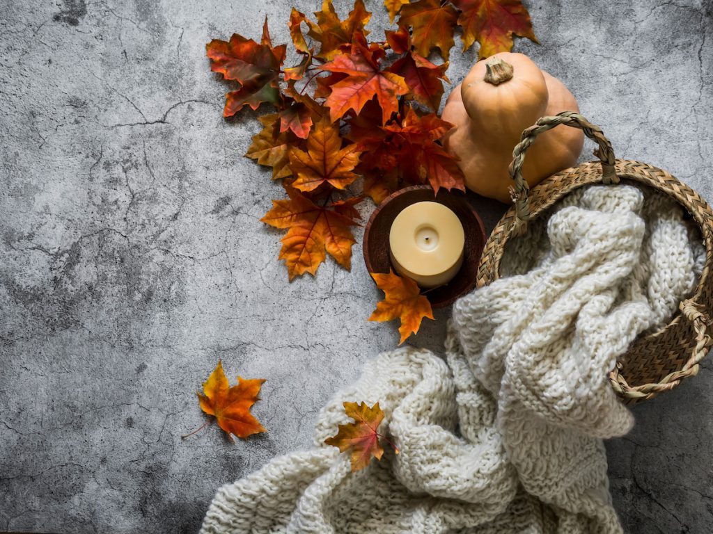 Autumn still life. Wicker straw basket with knitted blanket, bouquet of maple leaves, candle on a gray background