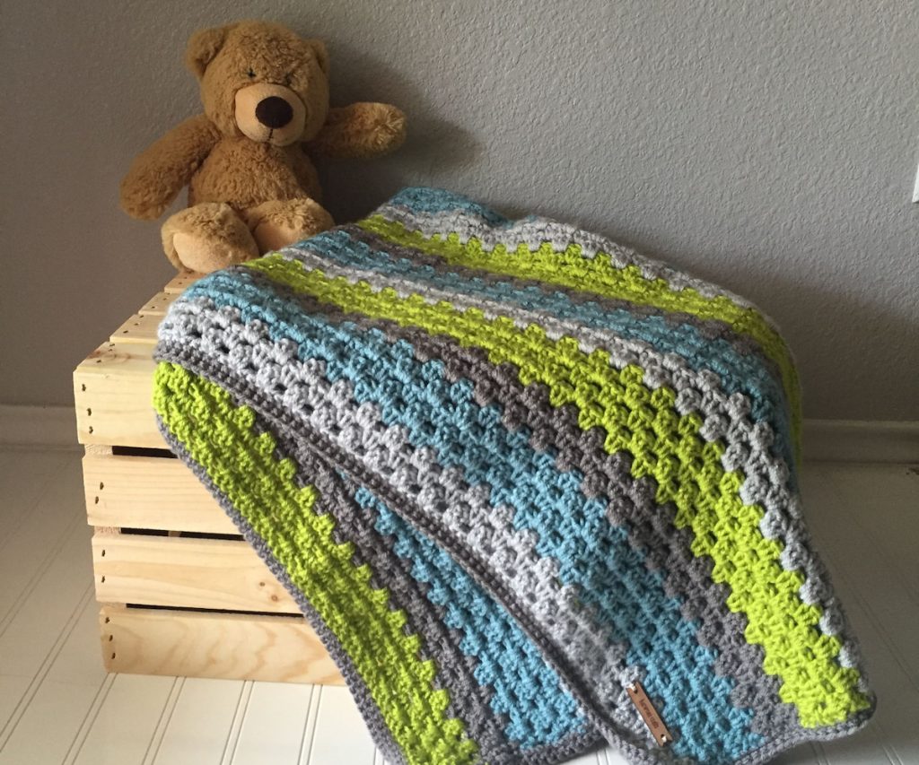 crochet granny stripe blanket with blues greens and grays sitting on a wooden crate with a brown teddy bear