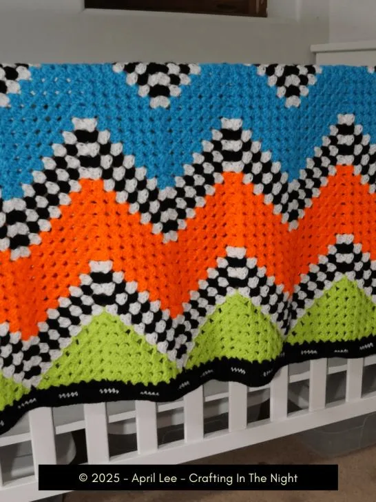 checkered chevron crochet blanket draped over crib. blanket has checkered sections alternating between solid colored stripes of neon green, orange, and neon blue